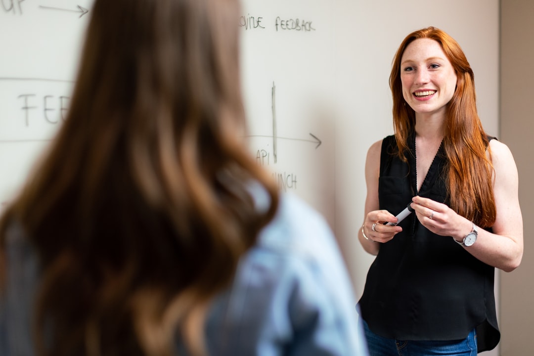 A teacher holding a marker stands at a whiteboard, explaining a math concept to a student.