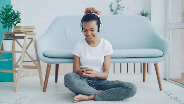 A teenage girl is sitting on the floor in a living room listening to music through headphones. Music can help improve memorization.