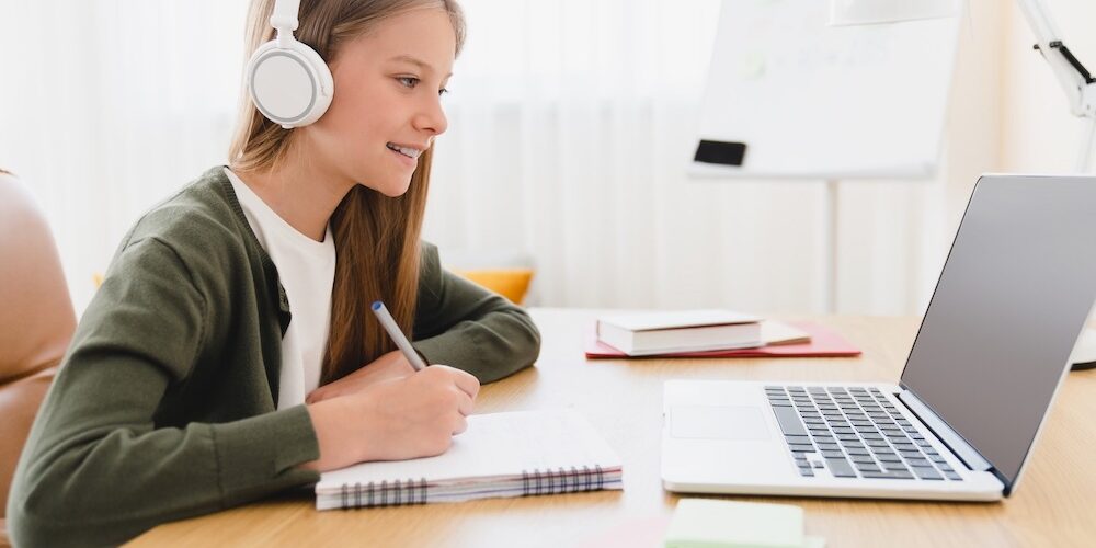 Distance remote education e-learning. Young caucasian schoolgirl student pupil doing homework studying from home using laptop and headphones, listening to online classes lessons.